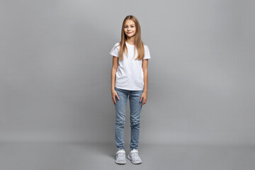 Full length portrait of happy teenage girl with long straight blonde hair standing on gray studio background. Smiling preteen child model in white t-shirt, jeans and sneakers posing confidently © Anna Zhukkova