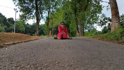Fototapeta premium Woman in Red Outfit Sitting on Road Surrounded by Trees on a Sunny Day, Nature Portrait