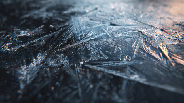 Detailed close-up of intricate frost patterns covering a dark surface, revealing delicate ice crystals beautifully illuminated by soft, cold light.