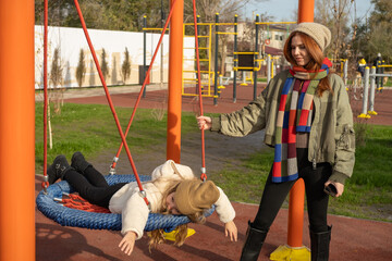 Mom and her daughter swinging on swing in autumn park. Happy family, child outdoors, weekend. Child girl, sits on swing smiles. Playground in fall.