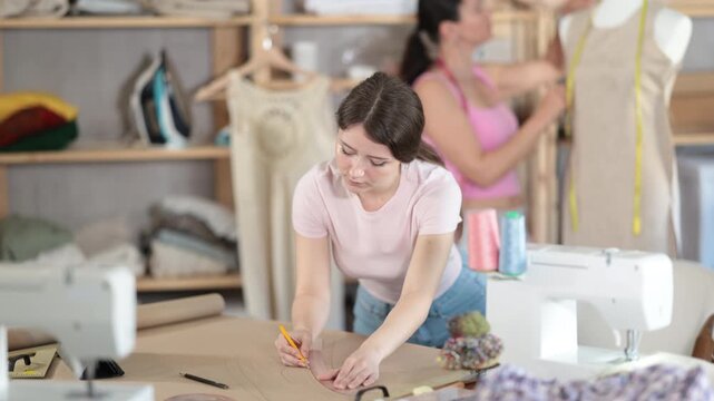  Using ruler and chalk, woman tailor draws dress diagram scheme on paper. Craftsman creates pattern for making dress. Companion worker in background with mannequin