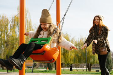 Mom and her daughter swinging on swing in autumn park. Happy family, child outdoors, weekend. Child girl, sits on swing smiles. Playground in fall.
