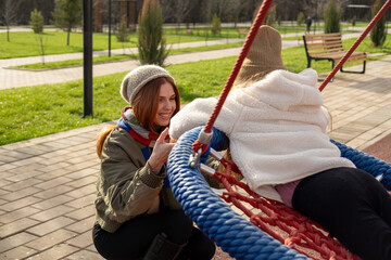 Mom and her daughter swinging on swing in autumn park. Happy family, child outdoors, weekend. Child girl, sits on swing smiles. Playground in fall.