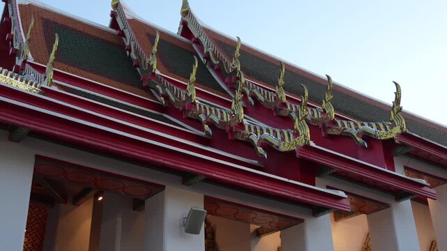 Ornate Wat Pho roof in Bangkok with vibrant red and green tiles, gilded chofahs and intricate decorative details against a clear sky, reflecting Thai spiritual heritage, slow motion reveal