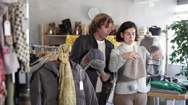 Focused european woman buyer looking hat for winter season and man helping choose right size in large boutique