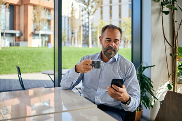 Businessman In Cozy Cafe Using Smartphone While Sipping Coffee Near Bright Window
