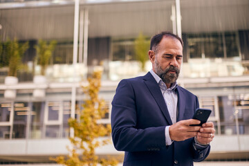 Professional Man in Navy Suit Using Smartphone Outdoors Near Modern Building for Business