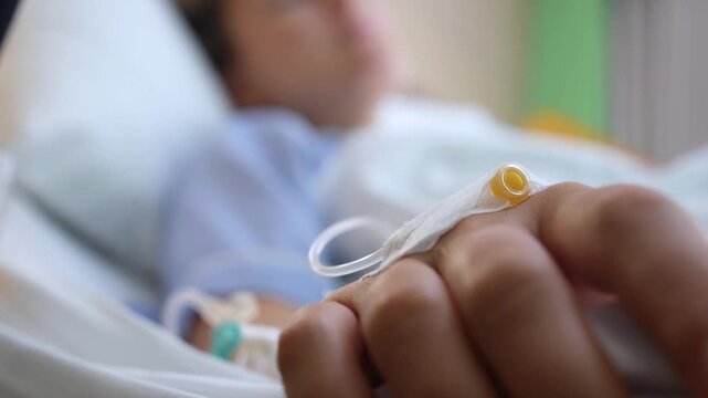 Close up of an unconscious patient's hand with an intravenous catheter attached for treatment, resting in a hospital bed with the person's blurred face visible in the background
