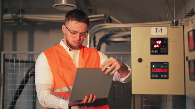 Young engineer wearing glasses and safety vest opens laptop, looking concerned while working in server room. Expert meticulously checks system, ensuring smooth operation with diligent focus.