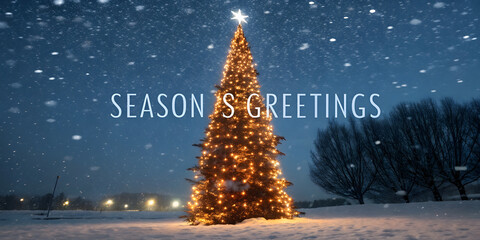 Christmas background image showing glowing outdoor Christmas tree covered with golden lights amid heavy snowfall under dark blue winter sky.
