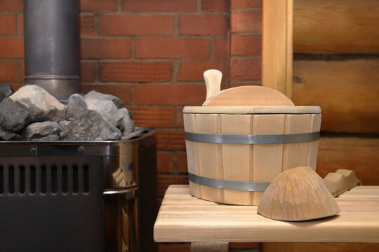 A wooden vat and ladle stand on a plank bench next to a stove with stones, against the background of a brick wall in a steam room. 