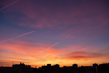 A vibrant sunset over a city skyline. The sky displays shades of purple, pink, and orange with scattered clouds and contrails.
