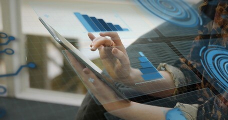 Tapping woman wearing patterned long-sleeve top holding tablet on sofa by window, with HUD charts