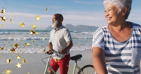 Riding mature couple wearing casual clothes cycling along sandy beach, with bicycles and gold stars