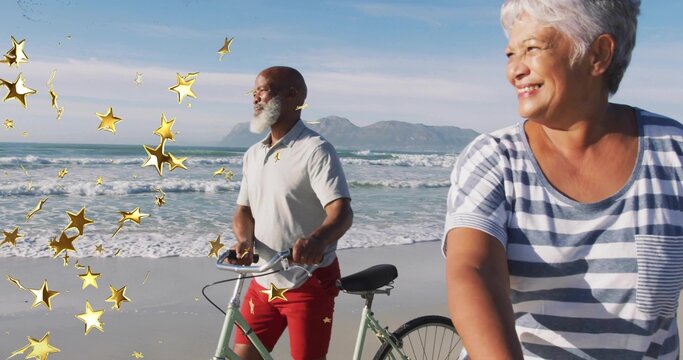 Riding mature couple wearing casual clothes cycling along sandy beach, with bicycles and gold stars