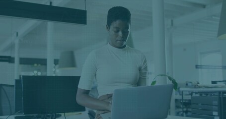 Standing woman wearing short-sleeve top checking silver laptop at open-plan workspace, with plant