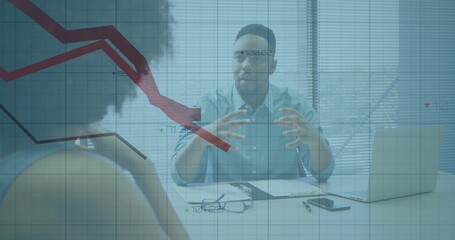 Talking man in blue shirt gesturing and explaining at office desk, with laptop and financial chart