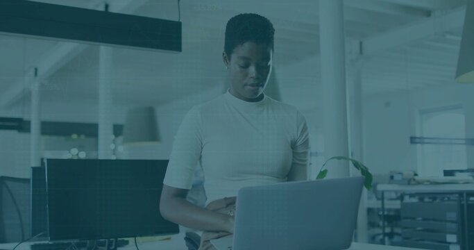 Standing woman wearing short-sleeve top checking silver laptop at open-plan workspace, with plant
