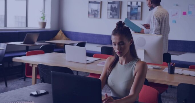 Working woman in sleeveless top with top knot typing on laptop at co-working space, with smartphone