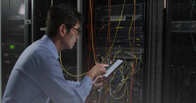 Repairing Korean tech in blue shirt leaning into server rack, using tablet tracing Ethernet cables - Powered by Adobe
