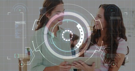 Holding tablet, two women wearing light blouse and striped top discussing data in office with HUD