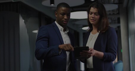 Pointing two colleagues reviewing tablet in modern corridor, with overhead lights, panels, blazers