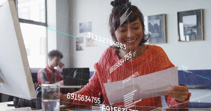 Reading woman in coral sweater smiling while holding paper at open office desk, data overlay