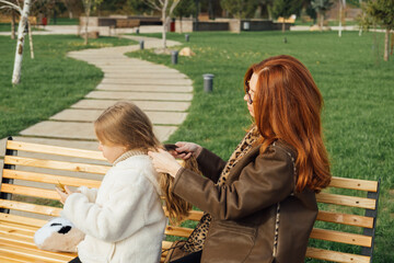 Mother braiding daughter hair on a park bench. Autumn setting, warm clothes, tender moment, outdoor.