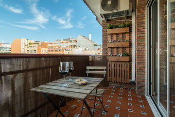 Obraz premium A cozy balcony in Barcelona with a wooden table and two glasses of wine. The view includes city buildings and a clear blue sky.