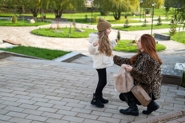 Mother and daughter walking hand-in-hand through a landscaped park in cool weather. Autumn vibes, warm clothes, peaceful moment.