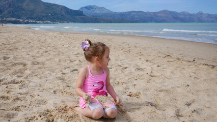 Little Girl In Pink Swimsuit Playing On Beach With Flower In Hair By Sea.