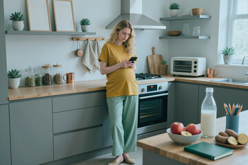 Pregnant young woman standing in modern kitchen using smartphone, holding belly with one hand, surrounded by fresh fruit and kitchen appliances, natural daylight entering window