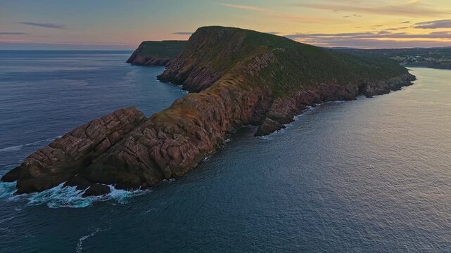 Wide aerial view reveals Torbay Point peninsula between two inlets, with rugged slopes, sheer cliffs and pastel sky stretching to the horizon.