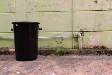 Black Trash Bin by Green Wall with Pipes and Dirty Ground in Urban Environment for Waste Management...