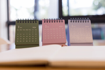 Colorful 2026 desk calendars showing January and February months on wooden table with blurred notebook ideal for planning and time management visuals.