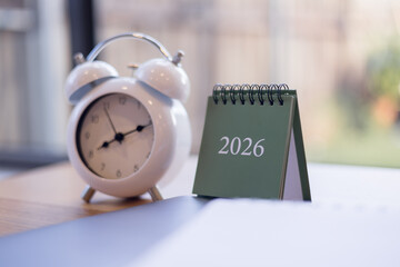 White retro alarm clock and green 2026 desk calendar on wooden table with soft daylight, perfect for time management, planning productivity themes.
