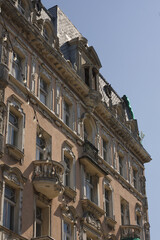 Detail of a richly decorated historic tenement house facade, showcasing ornate balconies, sculptural elements, and a classic mansard roof against a blue sky