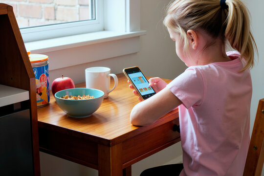 girl sitting at table using smartphone during breakfast, eating cereal with milk and apple nearby, focusing on screen, morning light coming through window