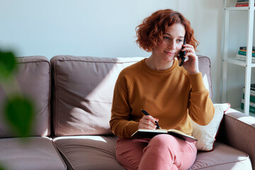 Young woman sitting on sofa talking on smartphone while writing in notebook, multitasking during remote medical consultation, focusing on healthcare communication