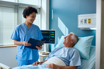 Black woman nurse standing beside hospital bed holding clipboard, smiling and talking to senior man patient lying in bed with medical monitor in background