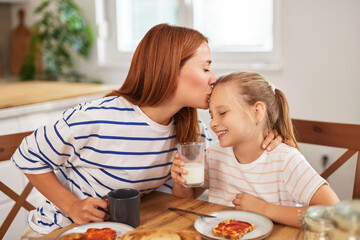 A mother affectionately kisses her smiling daughter while they share breakfast together at their cozy kitchen table, enjoying the warm atmosphere and each other’s company.