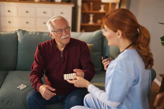 During the daytime, a female home healthcare nurse is sitting next to a senior man on a couch in his living room, handing him a blister pack of medication and monitoring his health.