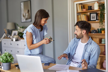 Obraz premium A woman sits on a desk, holding a coffee cup, engaging in conversation with a man who is seated in front of a laptop. They are in a cozy home office, effectively collaborating on their tasks.