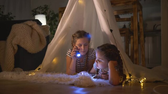 Girl reading with flashlight inside tent for cozy indoor play as sister and child imagine fort adventure under soft light at night blanket and string light surround creating warm calm atmosphere