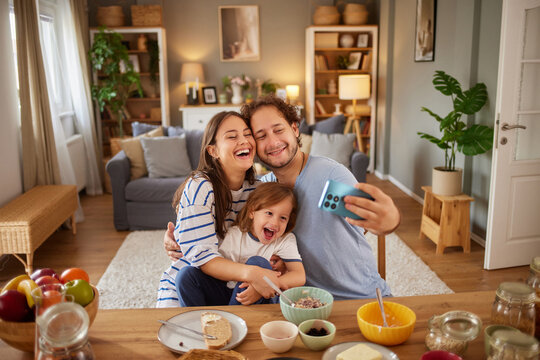A family shares a joyful afternoon moment in their cozy living room, surrounded by snacks. They laugh and take a selfie together, capturing happy memories. - Powered by Adobe