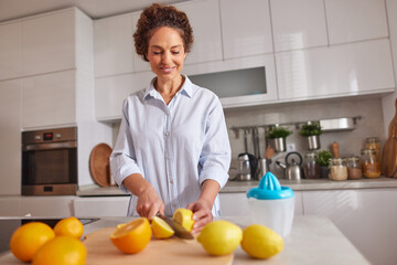 A cheerful woman is cutting lemons on a wooden board in a modern kitchen. Fresh fruits are scattered around as she prepares to make juice.
