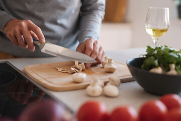 A person skillfully chops mushrooms on a wooden cutting board while surrounded by fresh vegetables and a glass of white wine in a bright kitchen.