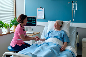 female nurse holding hand of senior woman lying in hospital bed, medical monitor displaying vital signs in background, providing compassionate care