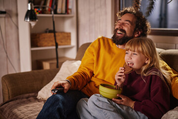 A man and a girl are sitting on a couch in a warm living room, laughing and watching a movie. They share snacks, creating a joyful atmosphere during their time together in the evening.