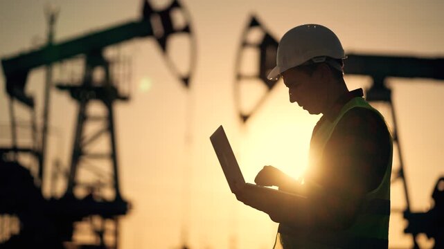 Worker inspects oil pumpjack silhouette at sunset using laptop while helmet wearing engineer monitors rig operation at industrial field with pumpjack and rig outlines in backlight during golden hour
